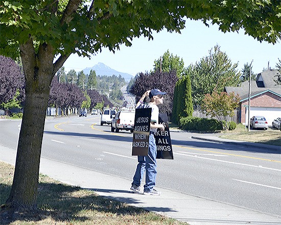 Gary Haga raises his arm and waves at motorists near Jennings Memorial Park while wearing his sandwich board sign to honor God. He plans to continue the evangelism as long as he can