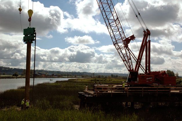 Construction crews steady a 50-foot steel pile as a crane uses a pile driver to drive it into the soil of Ebey Slough. It will help support the temporary work platform from which crews will build the new Ebey Slough Bridge.