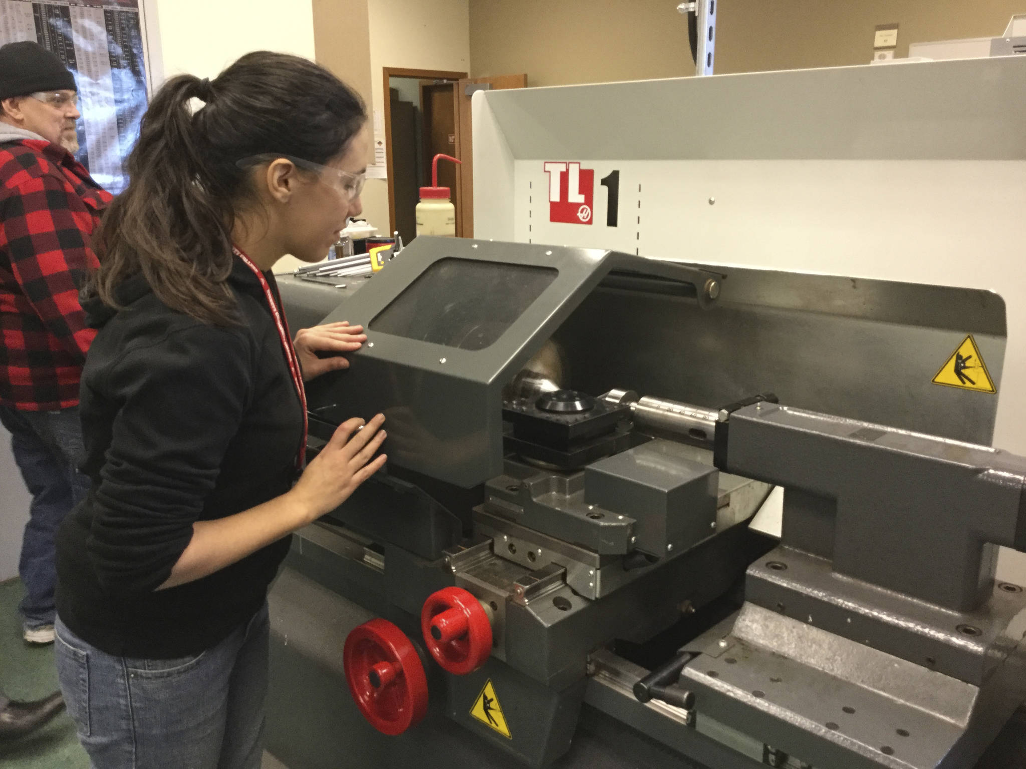 Instructional Technician Sophie Jessup, a former AMTEC student, operates a lathe that will produce a wood-grained writing pen, while teacher Jerry Becraft walks by in the machine shop at AMTEC North in Weston High School.